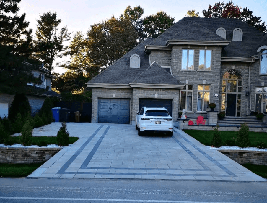 Entrée résidentielle en pavé uni gris avec motifs décoratifs foncés, double garage, murets paysagers et maison en pierre