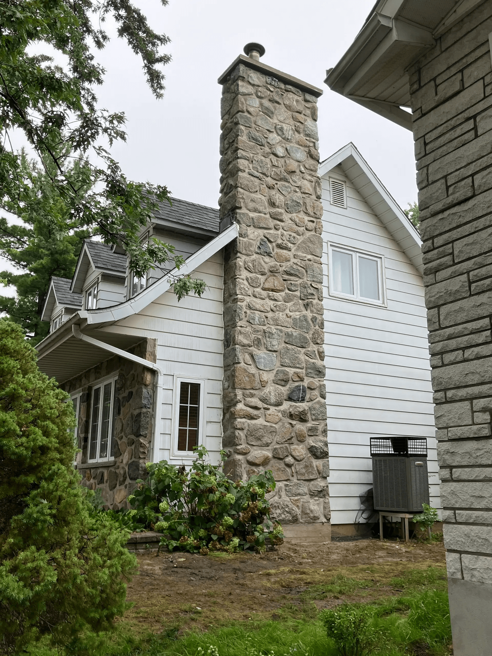 Grande cheminée extérieure en pierres naturelles sur une maison résidentielle avec revêtement blanc, toiture en bardeaux et aménagement paysager verdoyant 