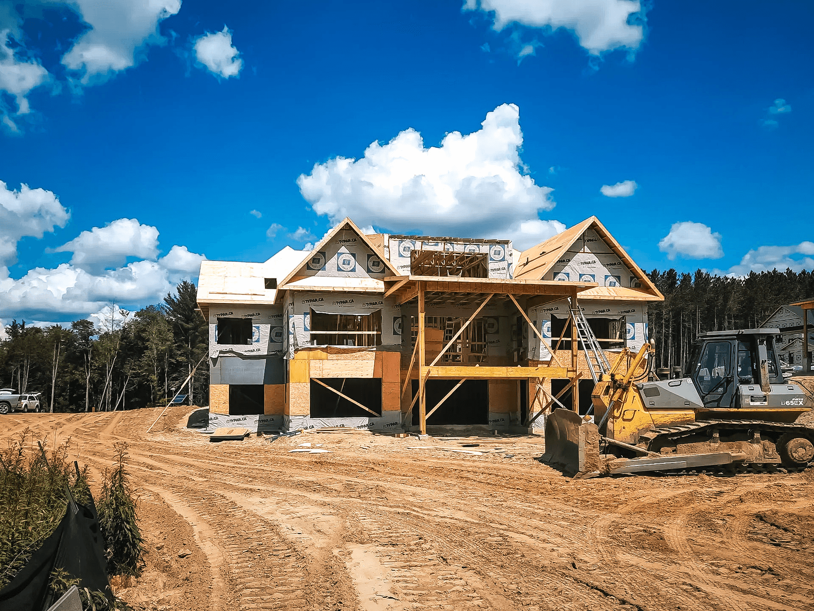 Maison à deux étages en construction sur terrain sablonneux avec pare-intempéries Tyvek, échafaudages en bois et machinerie lourde sur place.