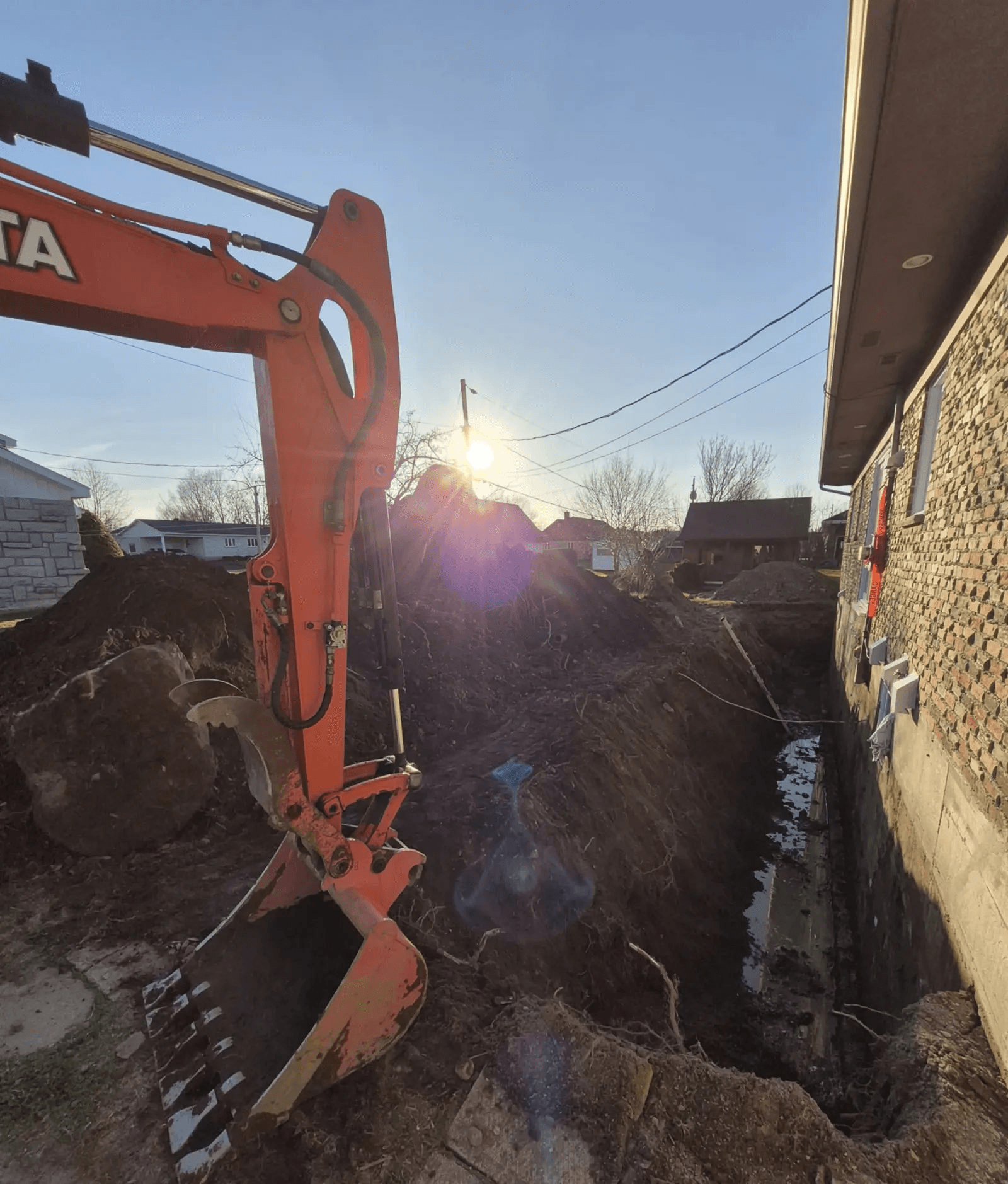 Travaux d'excavation autour d'une maison au coucher du soleil, à l'aide d'une pelle mécanique, pour la préparation des fondations ou l'installation d'un drain français.