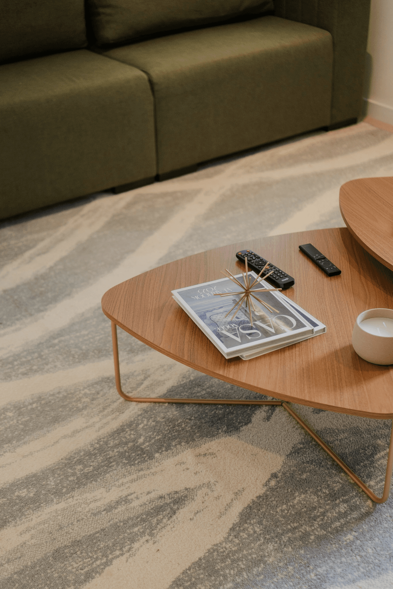 Salon moderne avec un canapé vert olive, un tapis abstrait gris et beige, et des tables basses en bois sur lesquelles sont posés des magazines et des télécommandes.