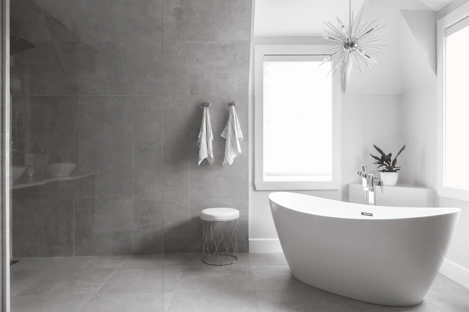 Salle de bain moderne et luxueuse avec baignoire autoportante blanche, douche vitrée à l'italienne, grands carreaux de céramique gris, robinetterie chromée, fenêtres lumineuses, luminaire suspendu contemporain et décoration minimaliste