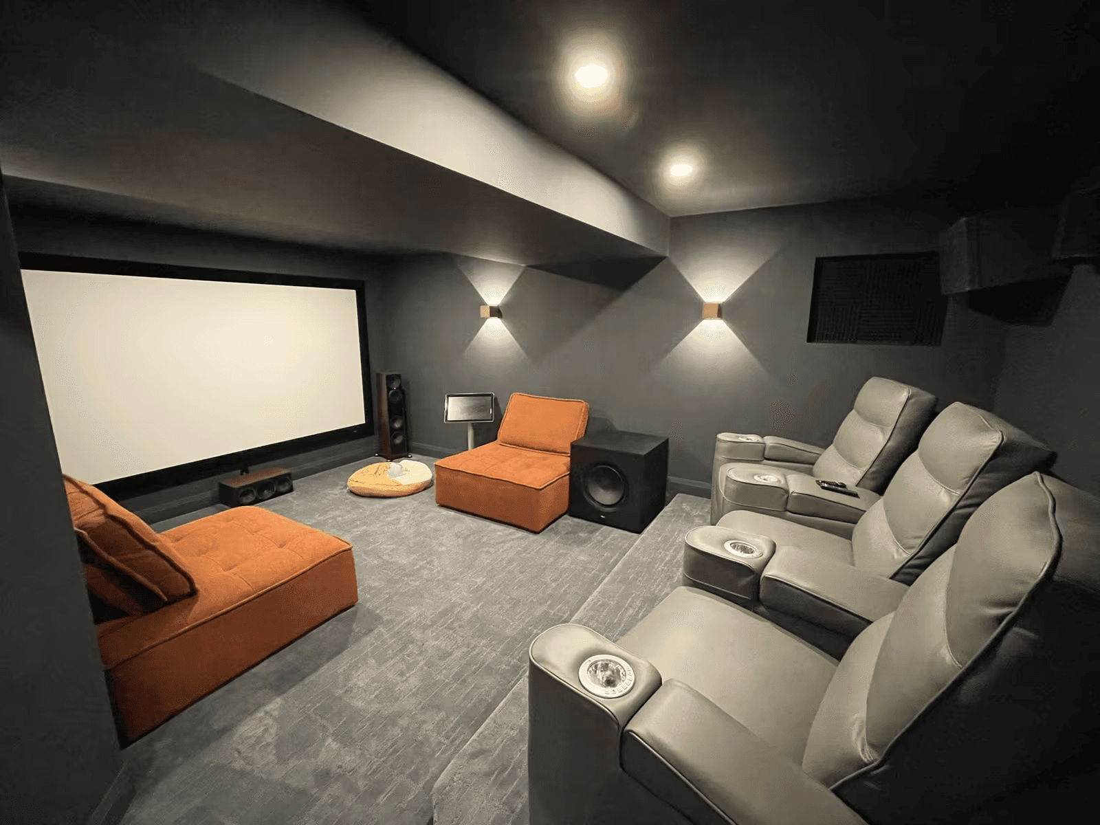 Cinéma maison sombre avec grand écran de projection, fauteuils inclinables gris, sièges lounge orange, système audio et éclairage mural moderne