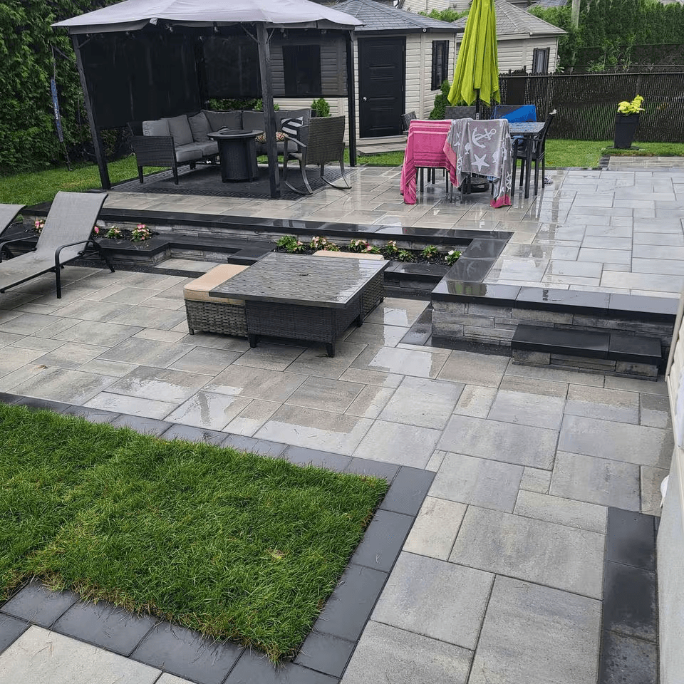 Aménagement paysager de cour arrière avec grande terrasse en pavé-uni gris, paliers surélevés, marches en pierre, bordures noires, jardinières intégrées, gazebo avec moustiquaires, mobilier extérieur, parasol vert et pelouse