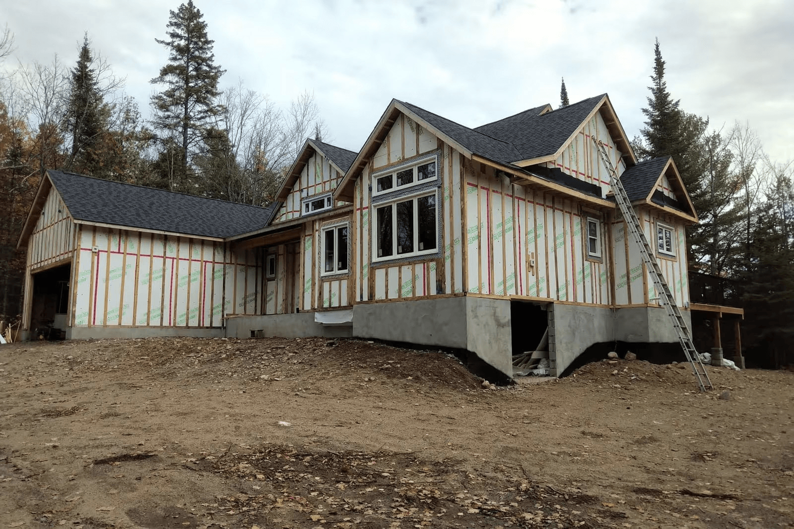 Maison en construction avec toiture en bardeaux, fenêtres installées, fondation en béton et façade protégée par une membrane pare-air avant la pose du revêtement extérieur.