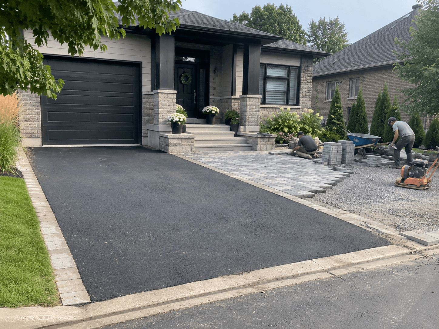 Travailleurs installant du pavé uni gris devant une maison résidentielle avec entrée asphaltée, bordures de pavés, gravier compacté et aménagement paysager