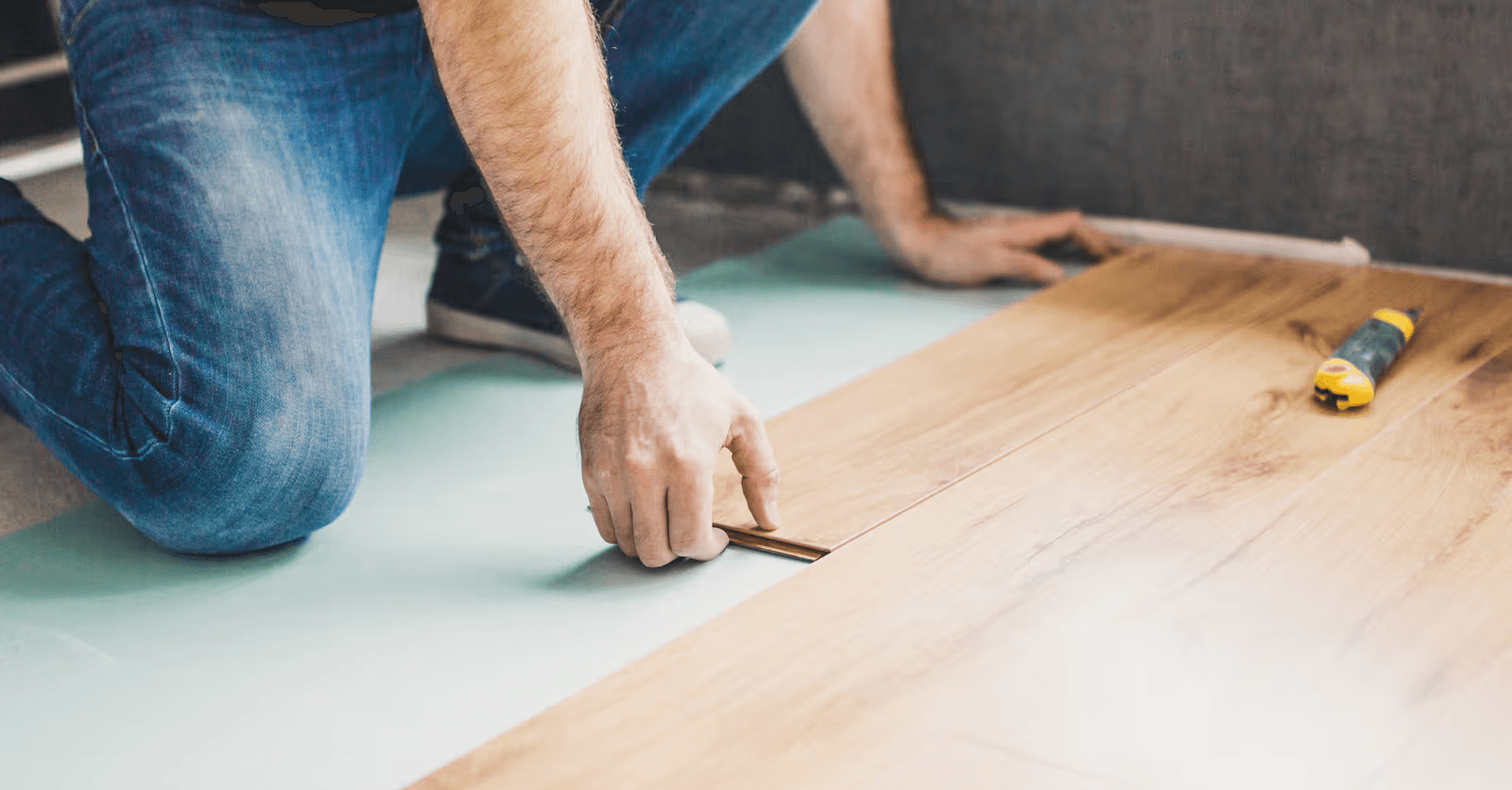 Installation d'un plancher flottant en lames de bois clair par un professionnel agenouillé, assemblant les planches sur une sous-couche isolante verte lors de travaux de rénovation intérieure résidentielle
