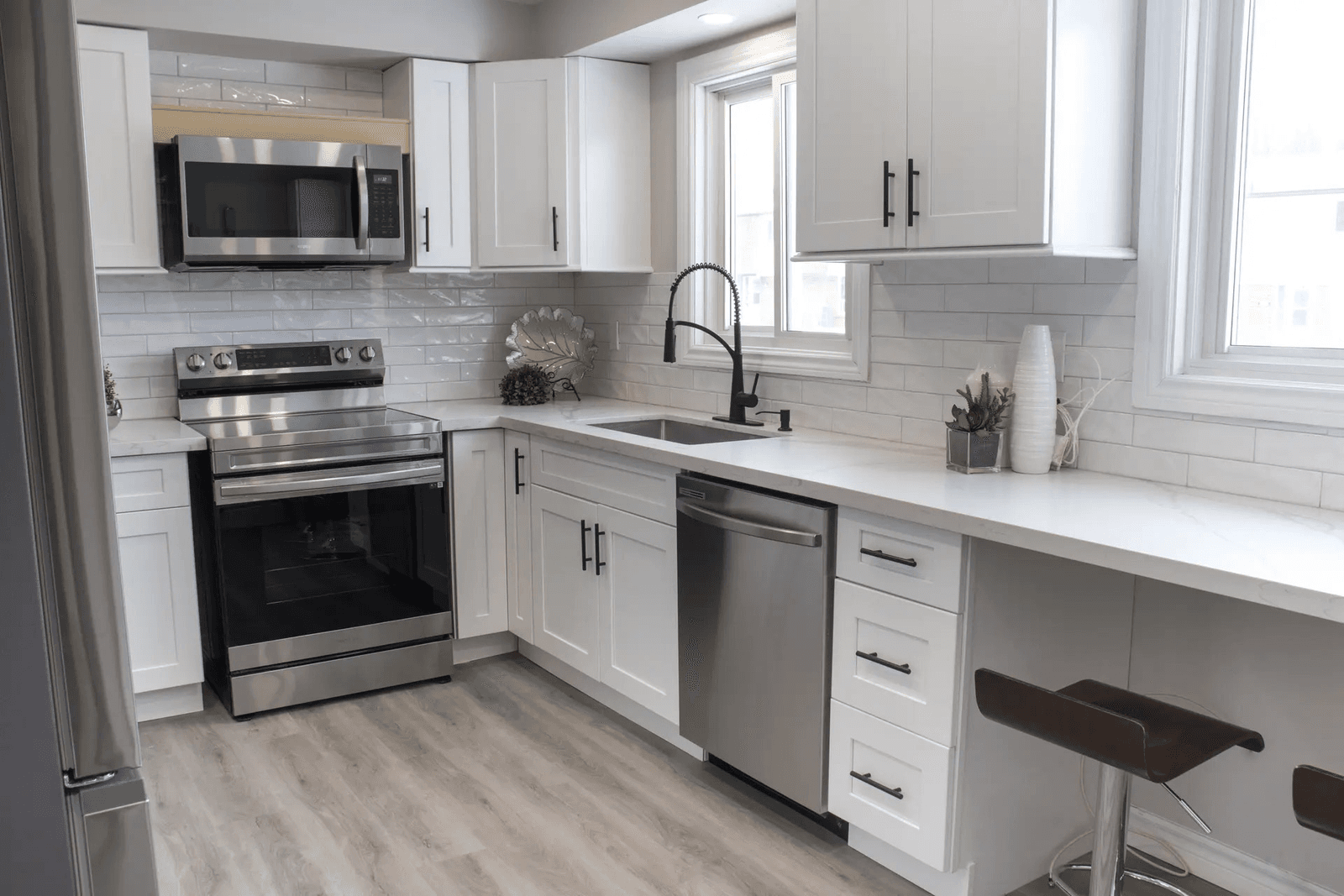 Cuisine moderne et lumineuse avec des placards blancs, des plans de travail clairs, une crédence en carrelage, des appareils électroménagers en acier inoxydable, un évier avec un robinet noir et un comptoir-bar avec des tabourets.

