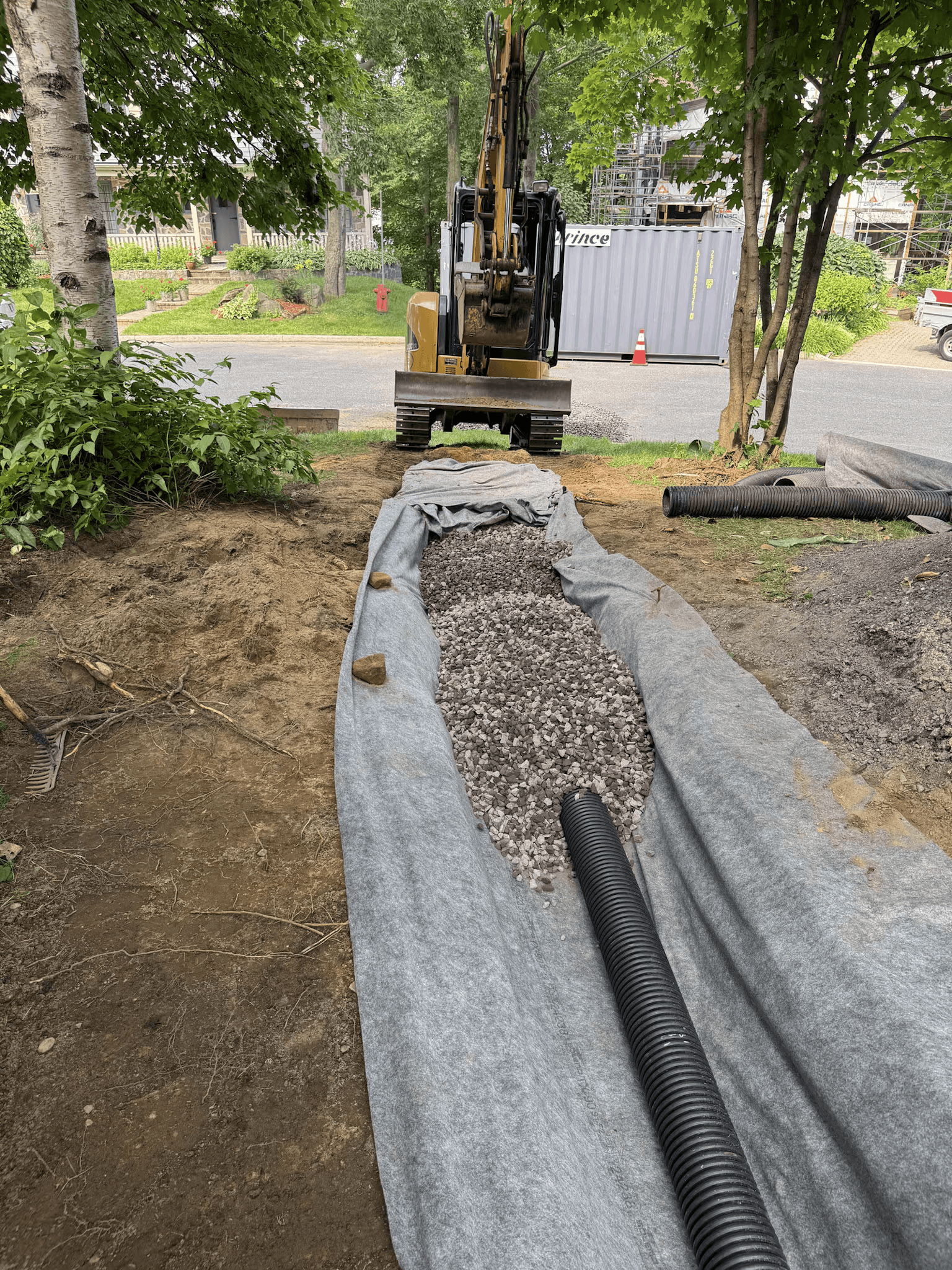 Installation d’un drain souterrain avec tuyau perforé, pierre concassée et membrane géotextile dans une tranchée résidentielle, avec excavatrice en arrière-plan.