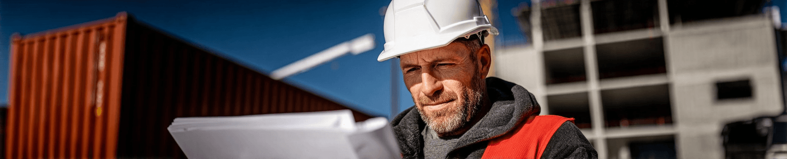 Entrepreneur en construction portant un casque blanc et un gilet de sécurité rouge, examinant des plans sur un chantier commercial extérieur