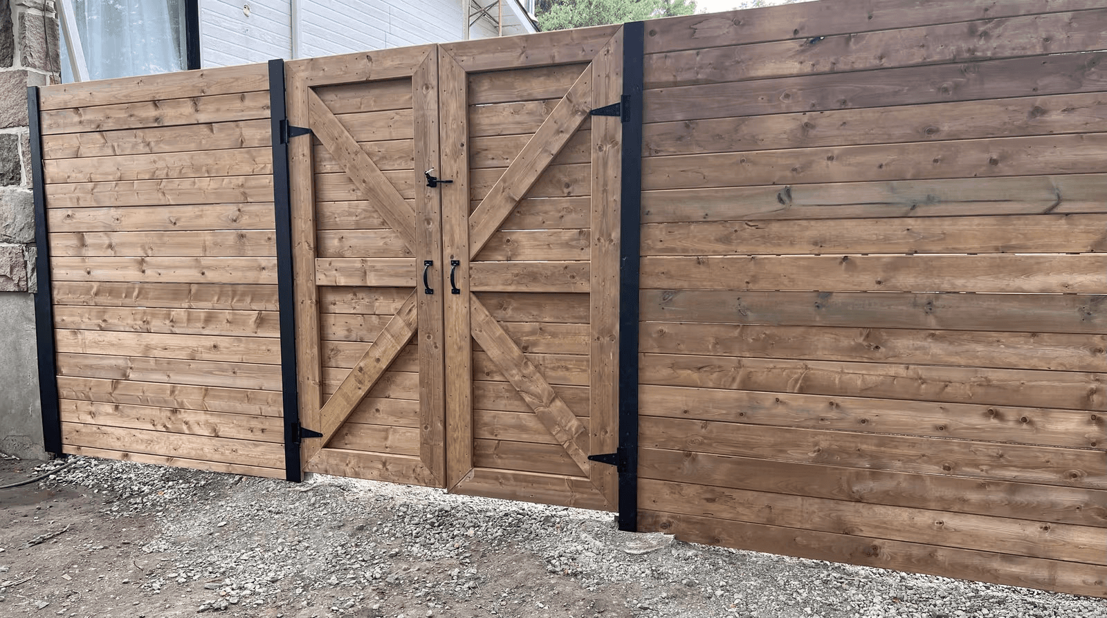 Clôture en bois à lattes horizontales avec double portail renforcé en diagonale, quincaillerie noire et poteaux métalliques, installée devant une maison résidentielle.