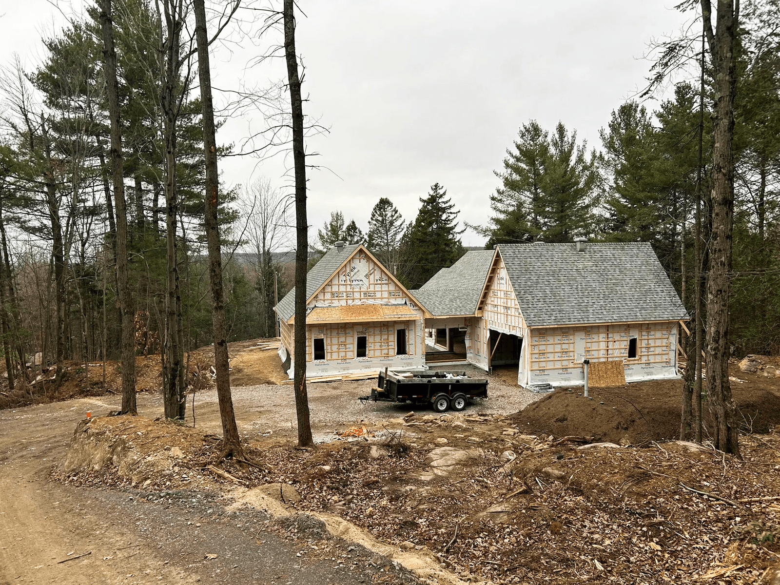 Maison en construction avec charpente et pare-intempéries visible, située en forêt sur un terrain en pente.