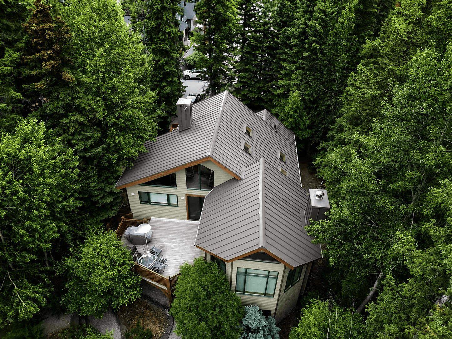 Vue aérienne d'une maison contemporaine entourée d'arbres, avec toiture en tôle grise à joints debout, grandes fenêtres et terrasse en bois.