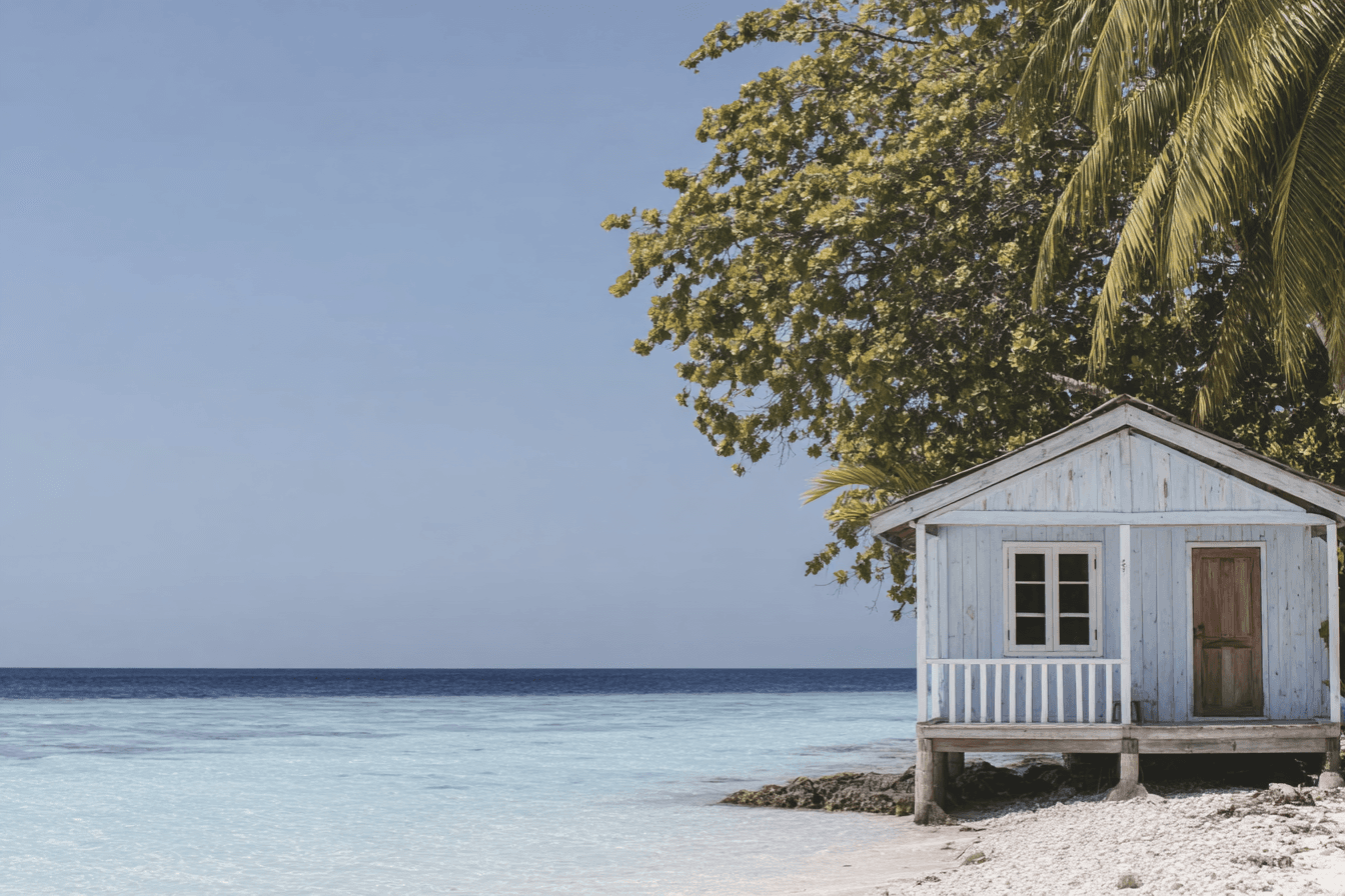Petite cabane blanche en bois sur pilotis au bord d’une plage tropicale, avec une mer turquoise, un ciel dégagé, des palmiers et une végétation luxuriante.