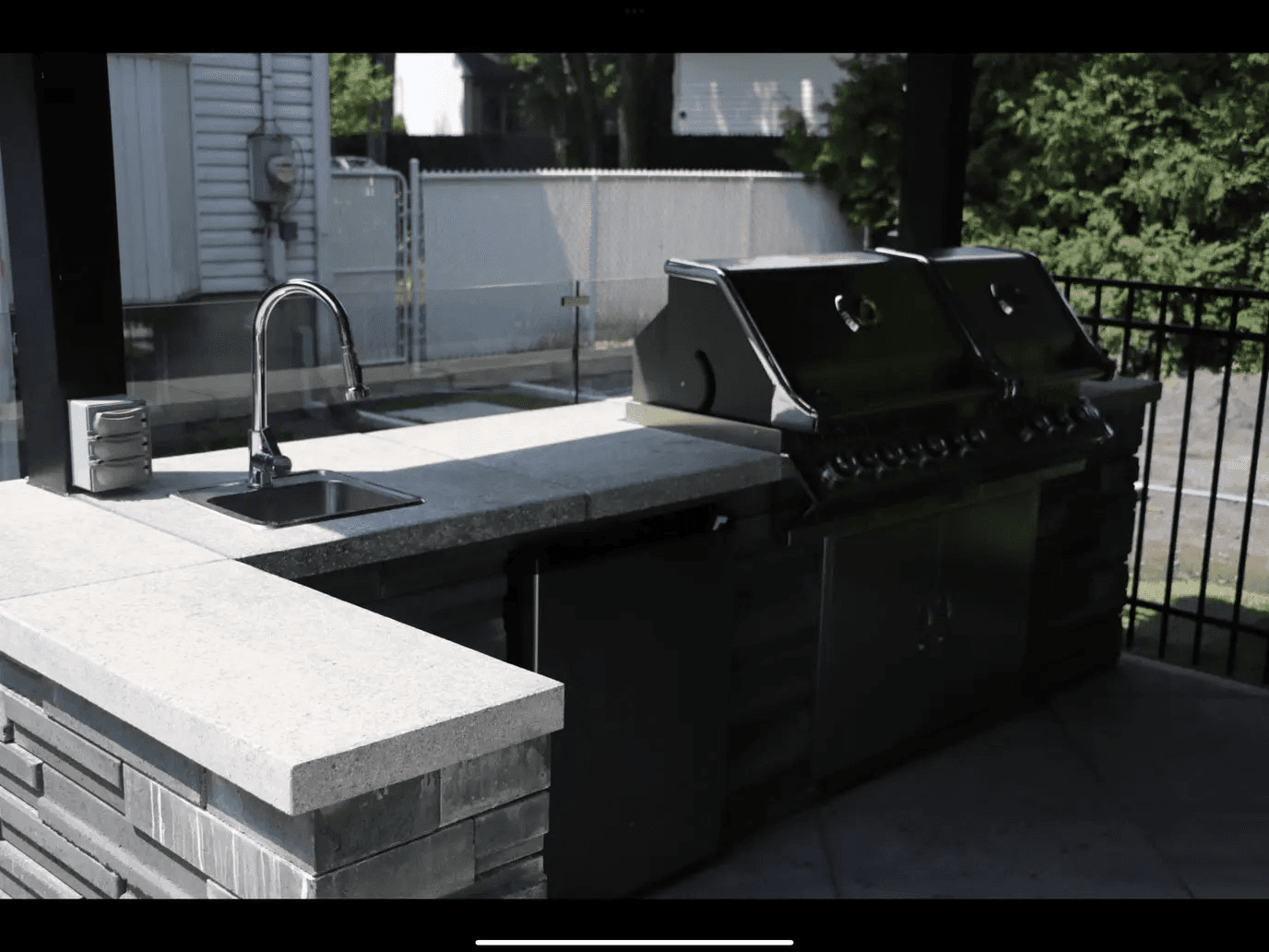 Cuisine d'été avec barbecue encastré, évier, plans de travail en pierre et rangements intégrés, sous une structure couverte.

