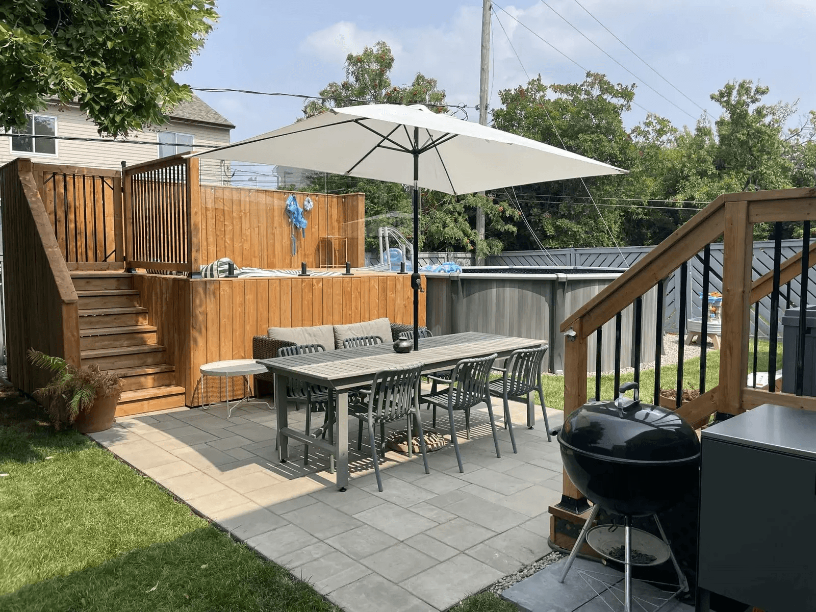 Aménagement de cour arrière résidentielle avec patio en dalles grises, table extérieure sous un grand parasol blanc, terrasse surélevée en bois avec escaliers et rampes noires, piscine hors terre, clôture d'intimité et barbecue