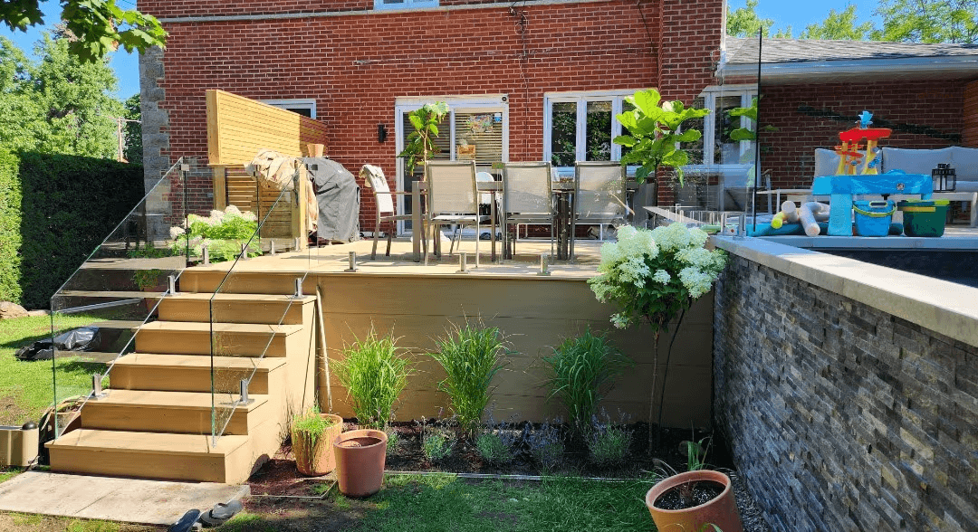 Terrasse résidentielle surélevée en bois avec escalier large, garde-corps en verre transparent, mobilier de jardin, panneaux d'intimité en bois et aménagement paysager avec graminées et hortensias, aménagée près d'une piscine avec muret en pierre grise et maison en brique rouge