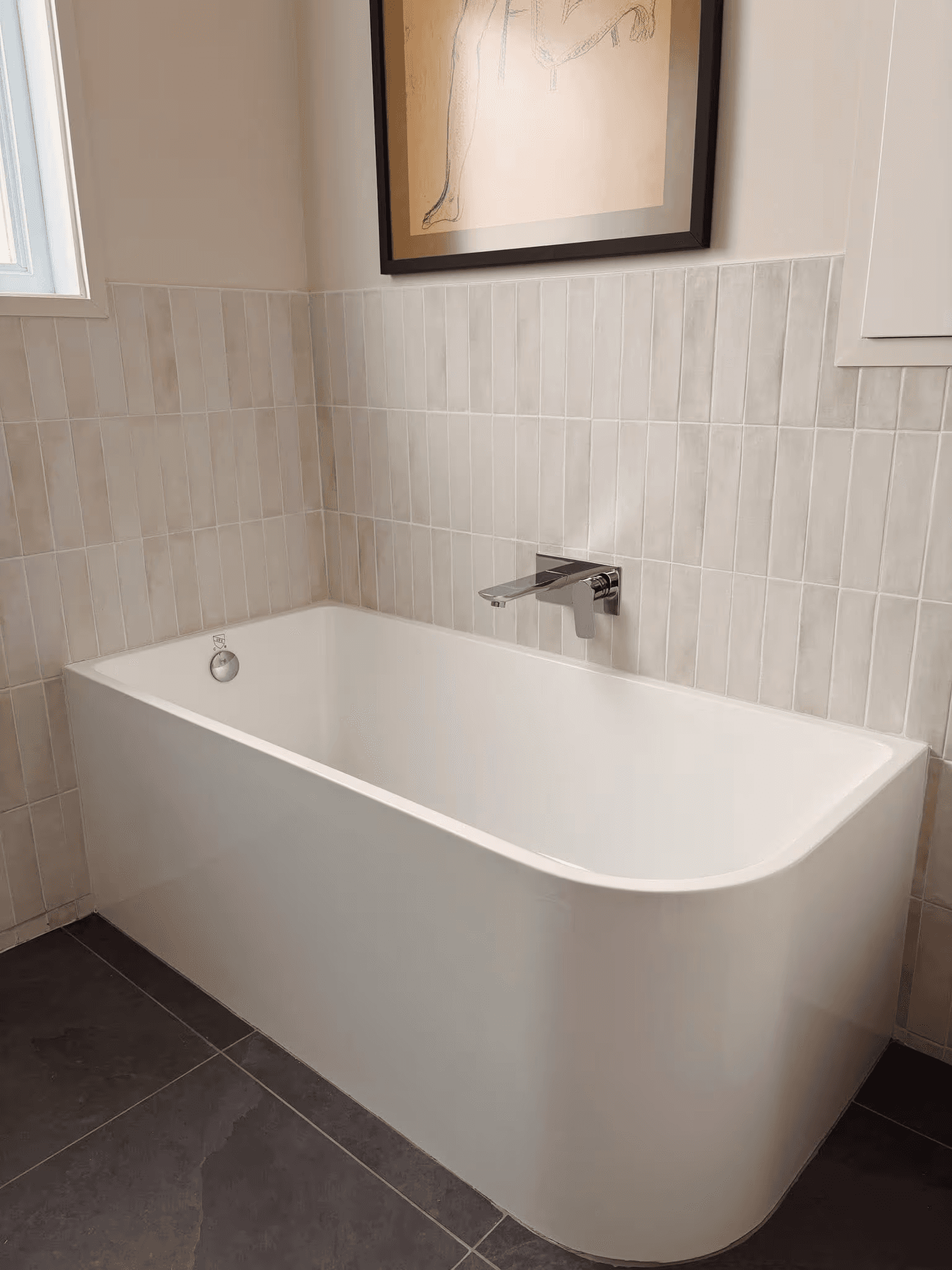 Salle de bain moderne avec un bain autoportant blanc de forme rectangulaire aux coins arrondis, une robinetterie murale chromée, des carreaux muraux beiges verticaux, un plancher de céramique gris foncé et un décor minimaliste.
