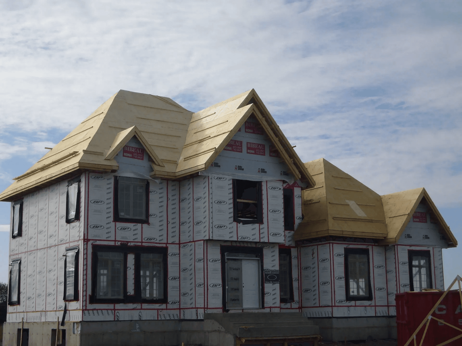 Maison à deux étages en construction avec membrane pare-air, toiture en contreplaqué, lucarnes, pignons et ouvertures de fenêtres installées.