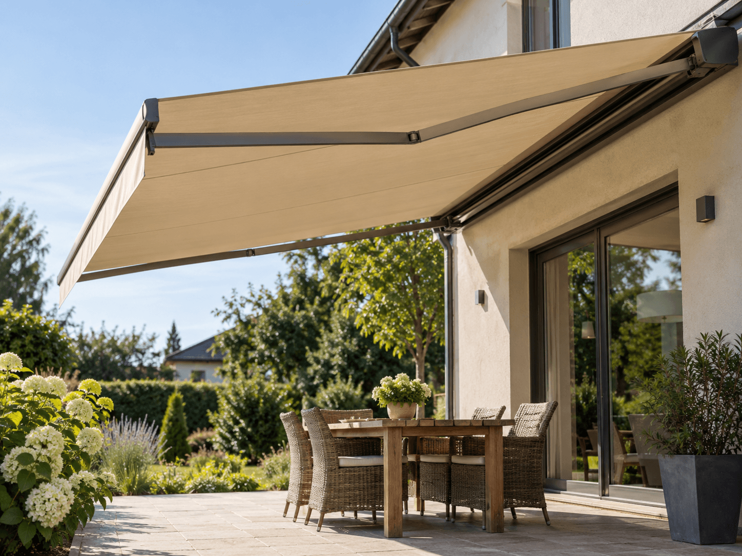 Auvent rétractable moderne en toile beige neuve avec bras articulés métalliques gris, installé sur une façade résidentielle en crépi pour créer une zone ombragée au-dessus d'une terrasse en dalles avec mobilier extérieur et jardin paysager