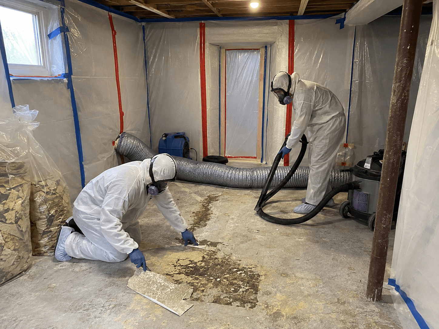 Travailleurs en combinaison de protection retirant un ancien revêtement de sol dans une zone confinée avec bâches de plastique, ventilation et aspirateur industriel /&nbsp;Workers in protective suits removing old flooring inside a contained work area with plastic sheeting, ventilation and an industrial vacuum