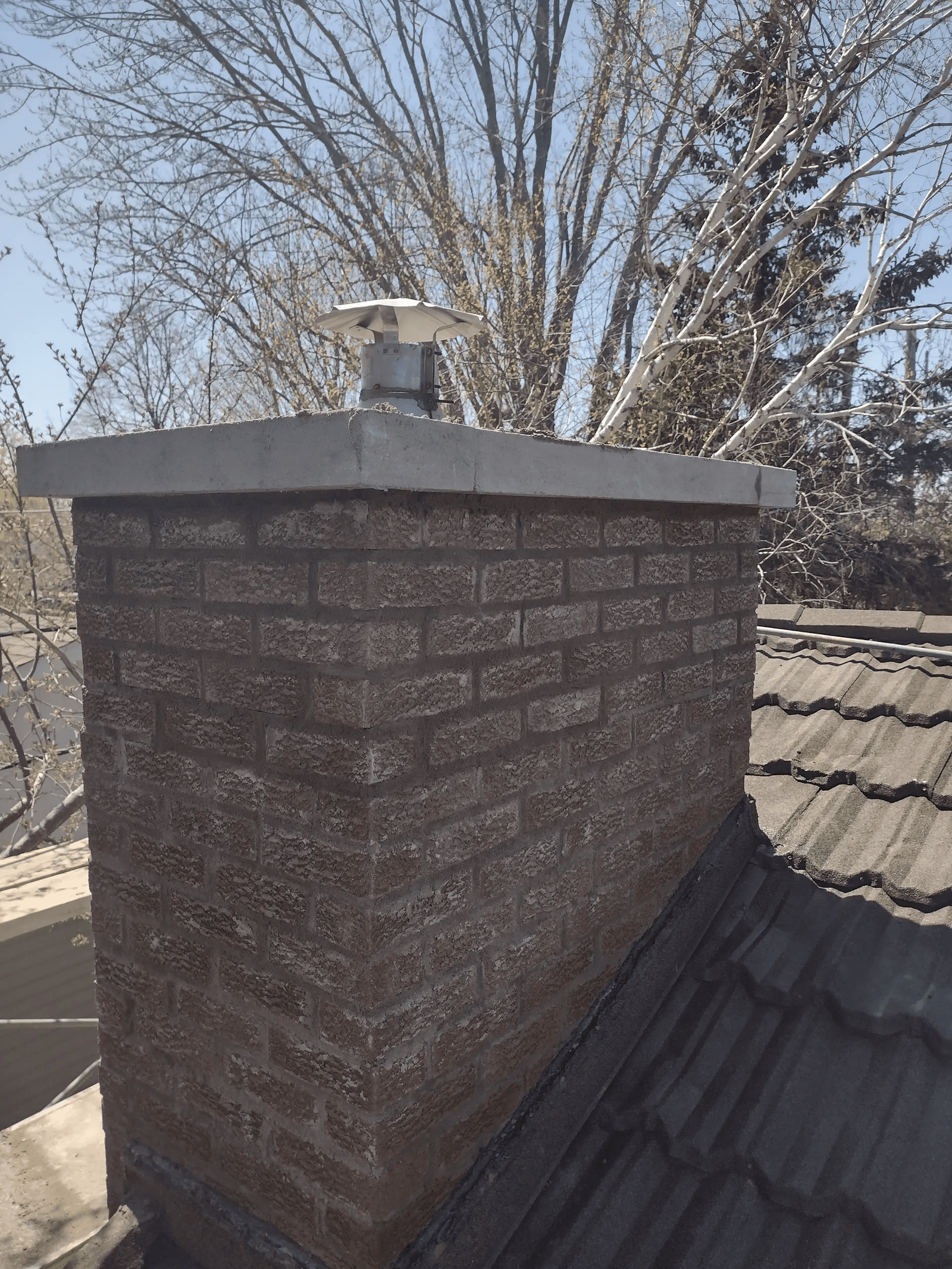 Cheminée extérieure en briques rouges avec couronnement en béton et chapeau métallique, installée sur une toiture résidentielle en bardeaux gris