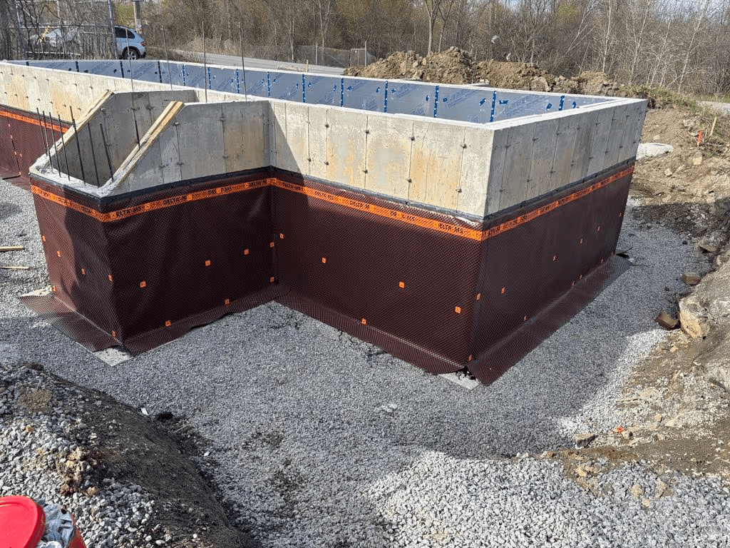 &nbsp;Fondation résidentielle en béton avec membrane drainante et imperméabilisante installée autour des murs, sur lit de gravier avant remblaiement