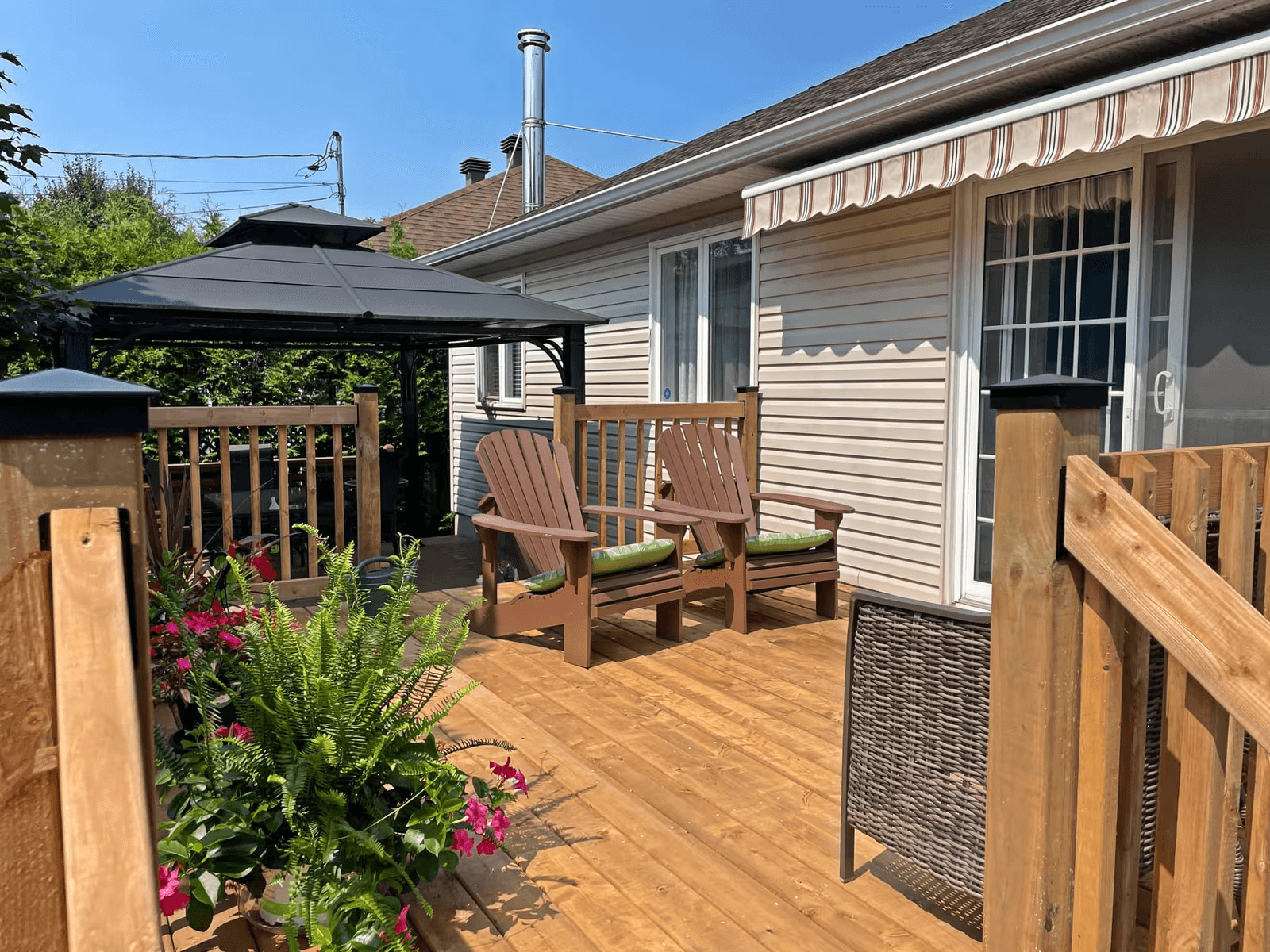 Terrasse résidentielle en bois naturel avec rampes et garde-corps, deux fauteuils Adirondack bruns, gazebo gris, auvent rétractable rayé, jardinières fleuries et revêtement extérieur beige de maison avec porte-patio vitrée