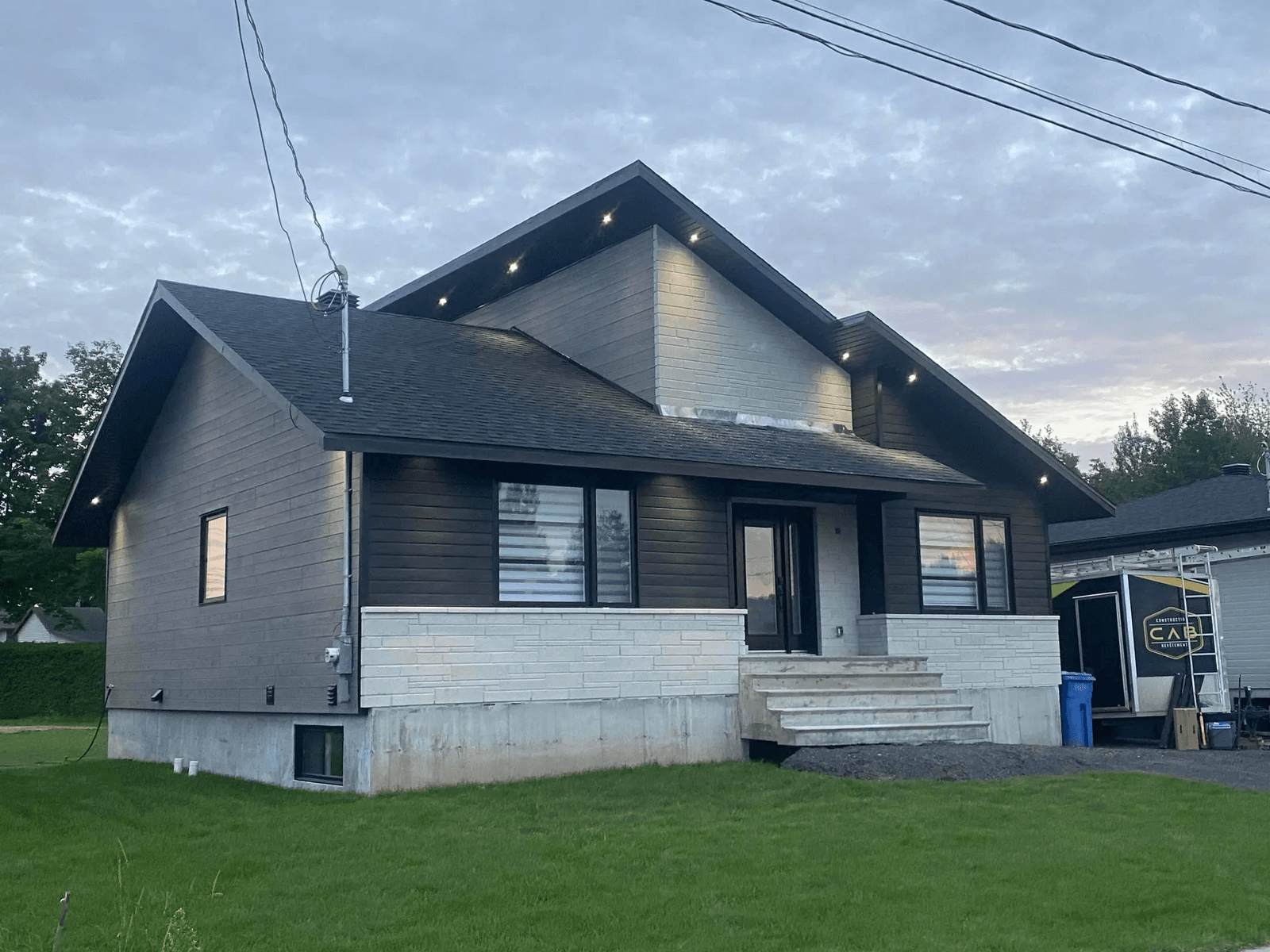 Une maison moderne avec un revêtement en bois foncé et de la pierre blanche sur la façade inférieure. Elle possède un toit à pignon avec des lumières encastrées sous les avant-toits, un escalier en béton menant à l’entrée principale et un garage visible sur la droite. La pelouse est bien entretenue, et le ciel est légèrement nuageux au crépuscule.&nbsp;