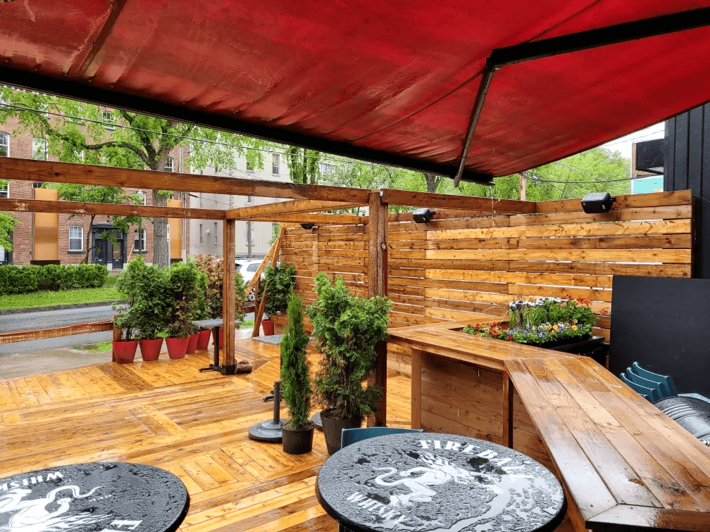 Terrasse extérieure commerciale en bois avec pergola, auvent rouge, clôtures en lattes horizontales, comptoir de bar intégré, plancher en bois, tables rondes et jardinières décoratives entourées de verdure