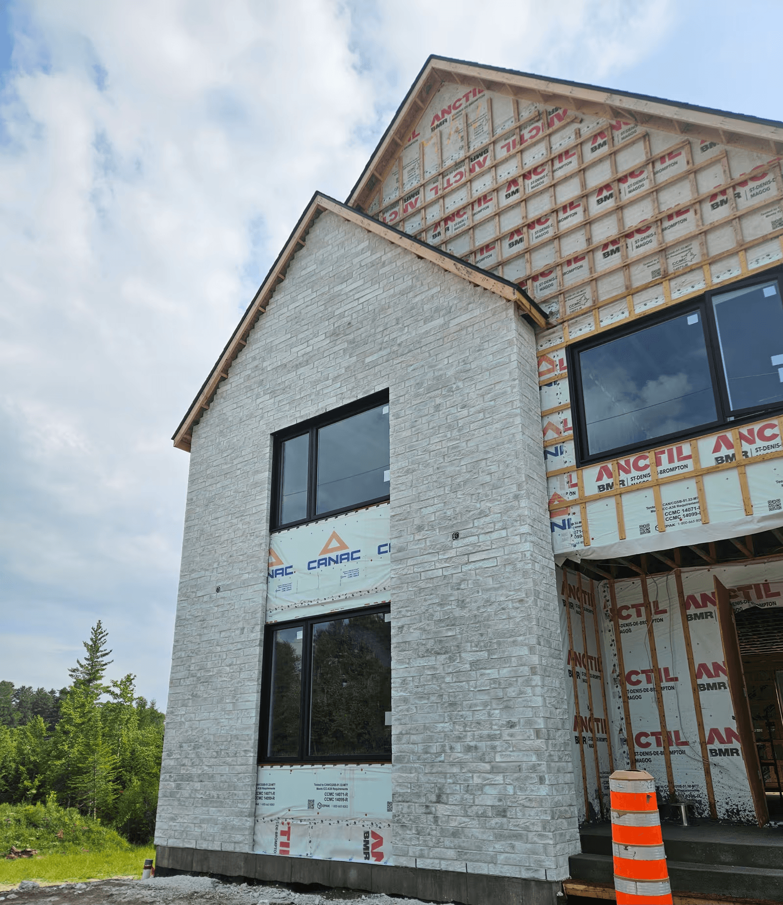 Maison neuve en construction avec façade en brique blanche partiellement installée, grandes fenêtres noires, membrane extérieure et fourrures de bois apparentes