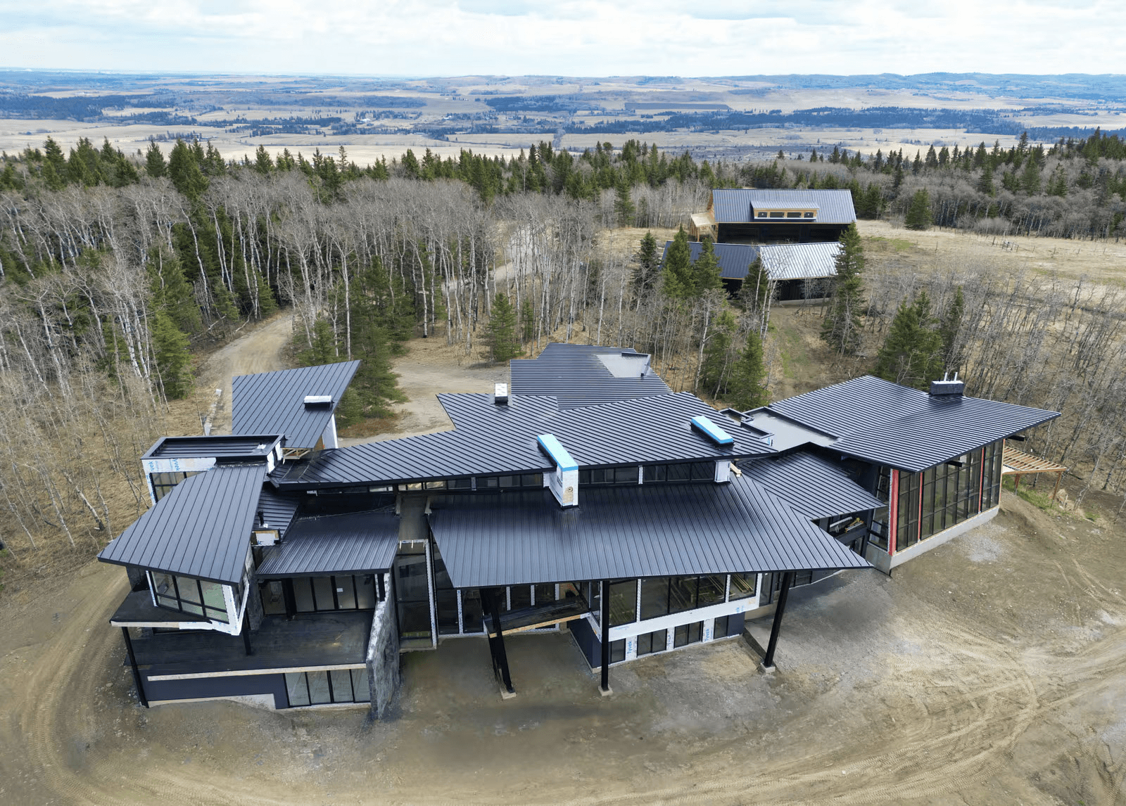 Vue aérienne d'une maison contemporaine en construction avec toiture métallique noire et fenêtres panoramiques, entourée d'une forêt.