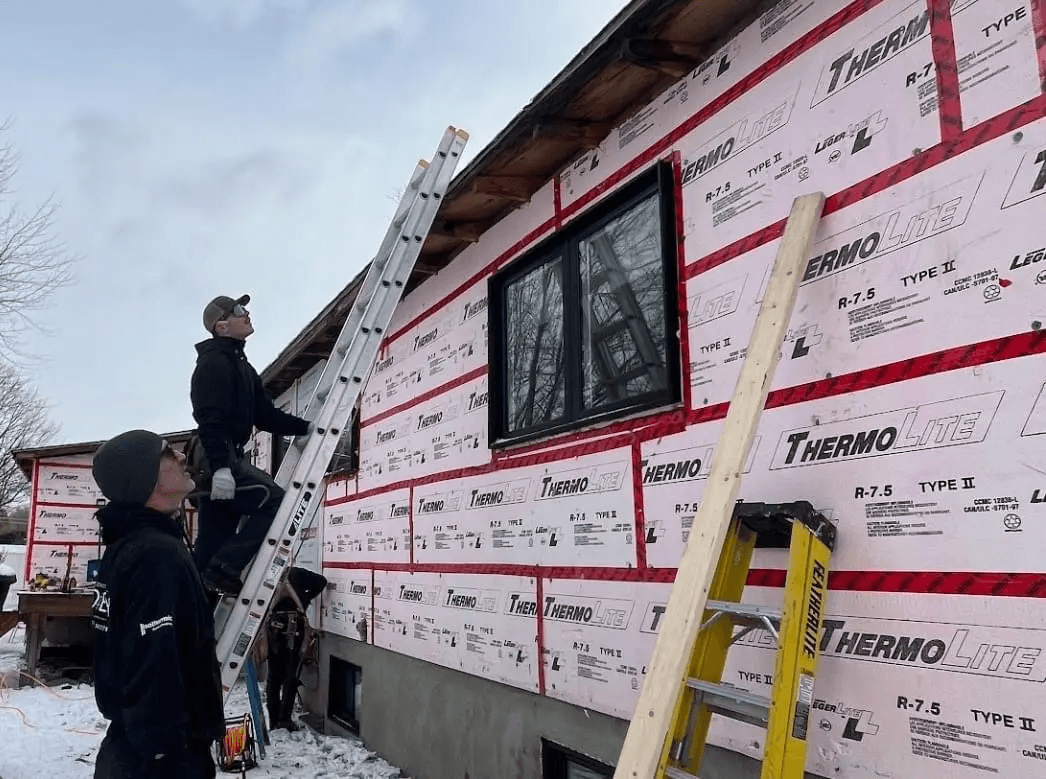 Travailleurs installant un revêtement extérieur isolant ThermoLite sur une maison en construction en hiver, avec échelles et outils de chantier.
