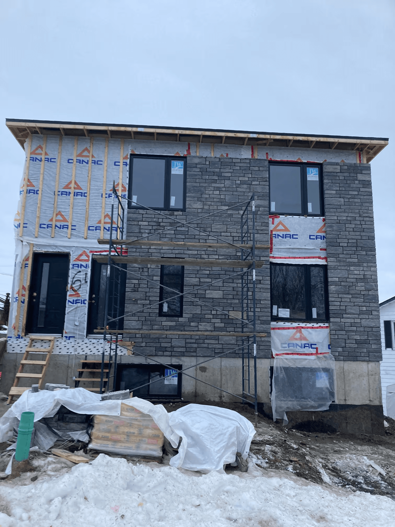 Construction d’une maison neuve à deux étages avec installation de maçonnerie extérieure en briques grises, échafaudage métallique, fenêtres noires et charpente de bois apparente