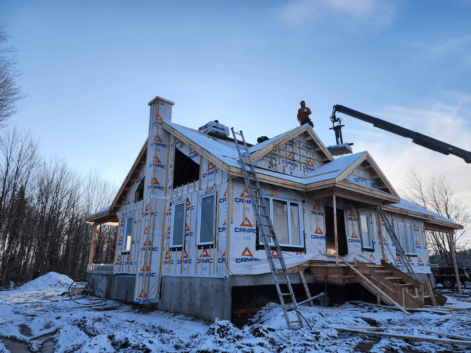 Maison en construction en hiver avec revêtement pare-intempéries CANAC, échafaudages, échelles, et ouvriers sur le toit sous un ciel bleu.