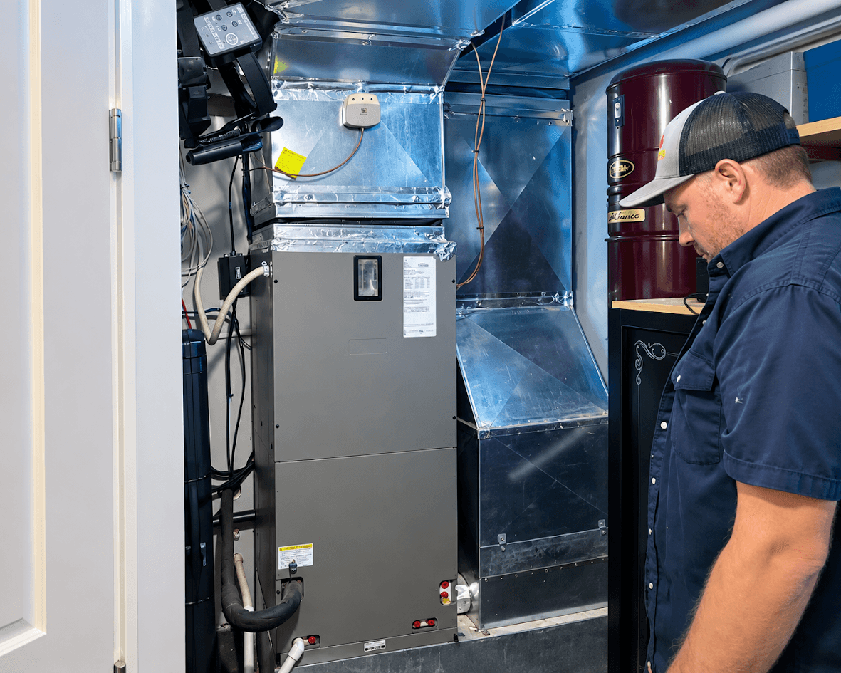 Technicien en chauffage et climatisation inspectant un système central avec unité mécanique, conduits de ventilation métalliques, tuyauterie et équipements techniques dans une salle mécanique résidentielle.