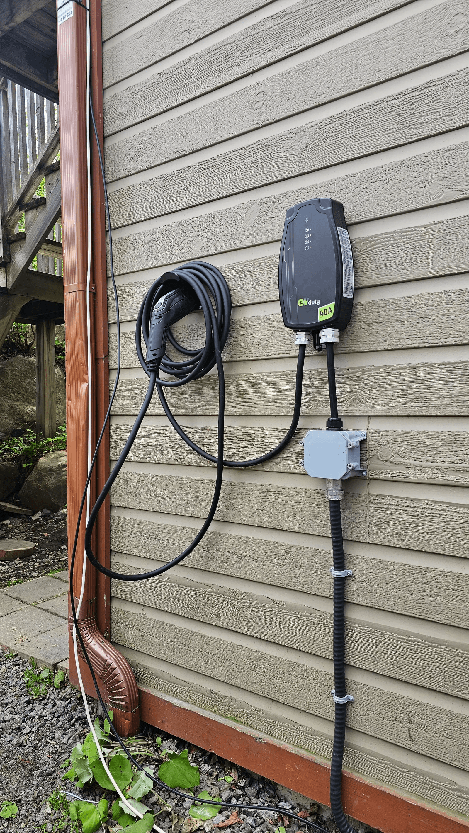 Borne de recharge électrique EVduty de 40 ampères installée sur le revêtement extérieur d'une maison, avec câble de recharge enroulé, connecteur et raccordement électrique apparent /&nbsp;EVduty 40-amp electric vehicle charging station installed on a house's exterior siding, featuring a coiled charging cable, connector and exposed electrical connections