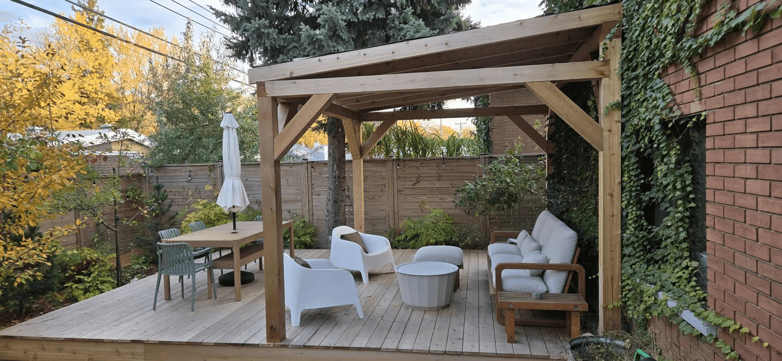Terrasse résidentielle en bois naturel avec pergola à toit incliné, poutres apparentes, poteaux robustes, salon extérieur avec canapé et fauteuils blancs, table à manger, clôture d'intimité en bois et aménagement paysager verdoyant près d'une maison en brique