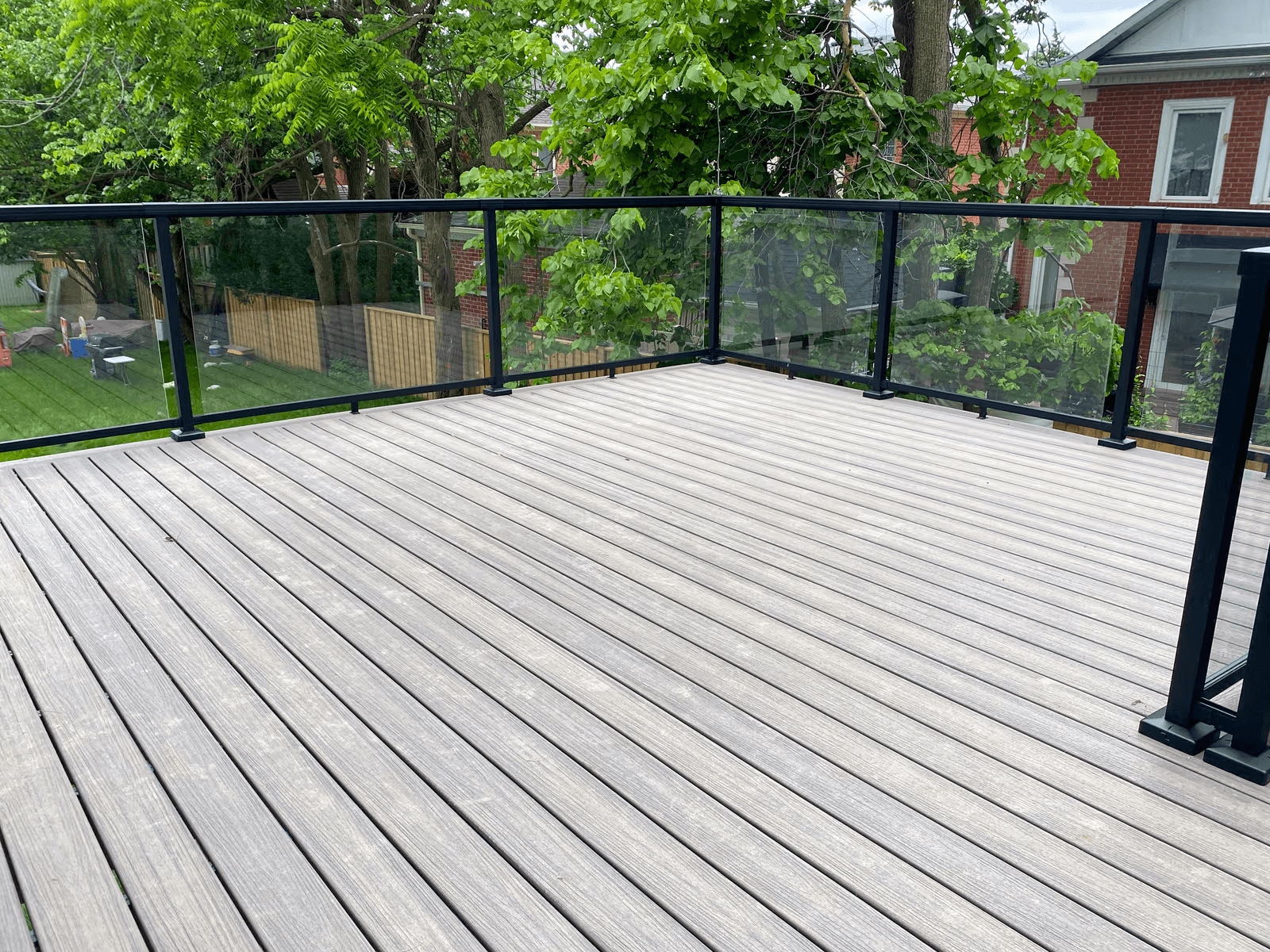 Grande terrasse résidentielle surélevée en planches de bois gris avec garde-corps modernes en verre transparent et structure en aluminium noir, offrant une vue dégagée sur une cour verdoyante avec arbres matures et maisons en brique