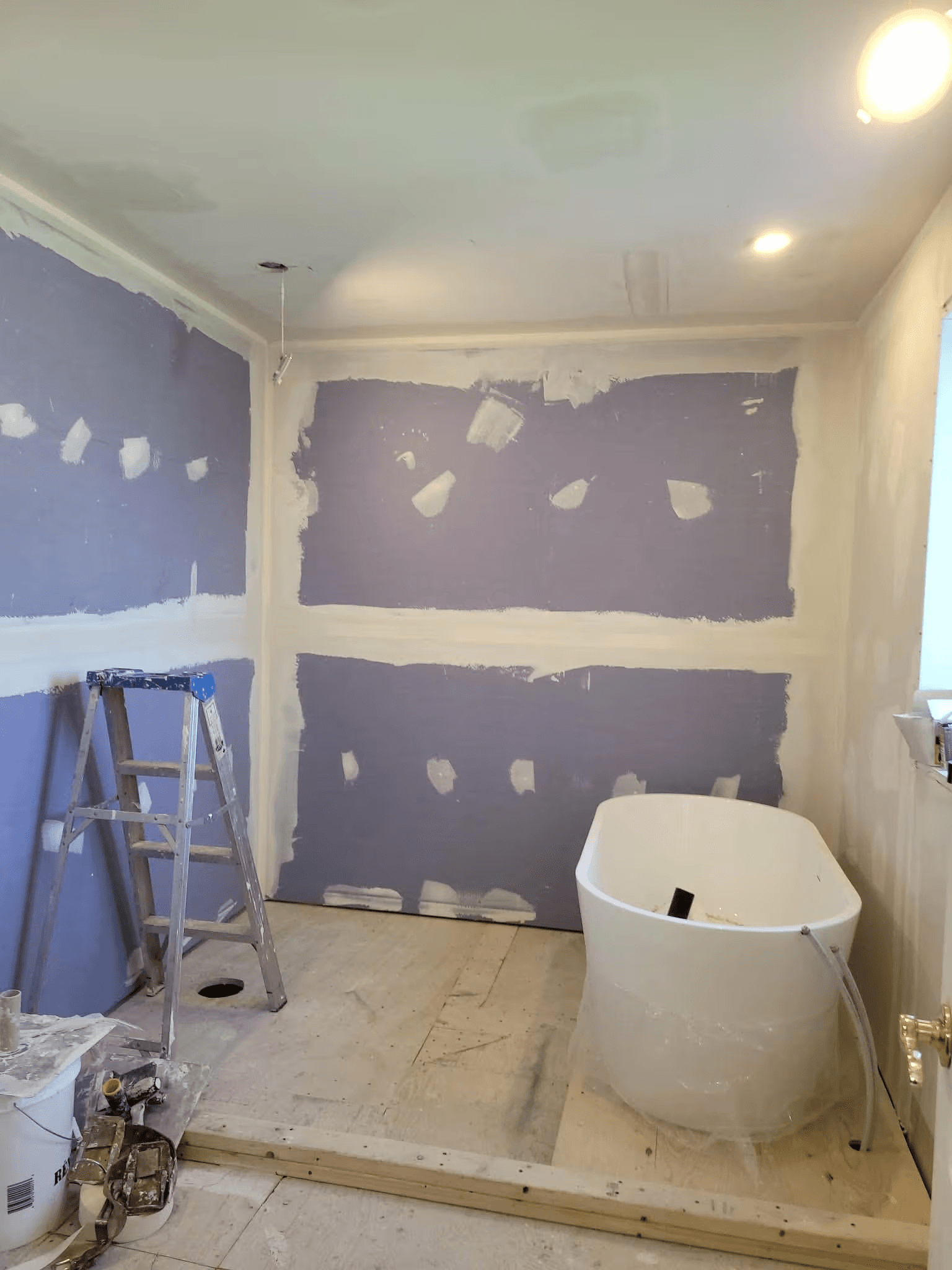 Rénovation d'une salle de bain en cours avec installation d'un bain autoportant blanc, panneaux de gypse violets, application de composé à joints, travaux au plafond, sous-plancher en contreplaqué et escabeau de chantier.