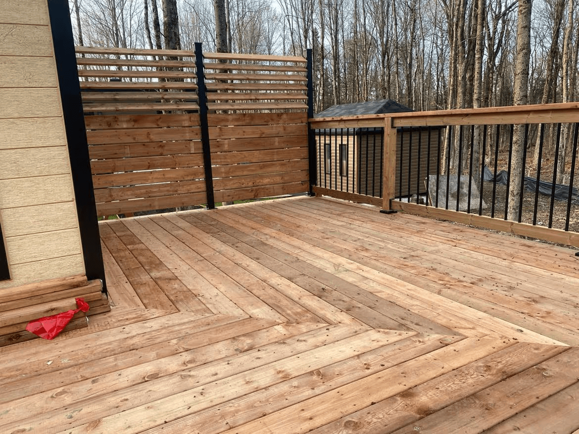 Grande terrasse résidentielle en bois naturel avec plancher en planches, garde-corps à barreaux métalliques noirs et panneaux d'intimité en lattes horizontales de bois, aménagée près d'une maison au revêtement beige et entourée d'arbres matures