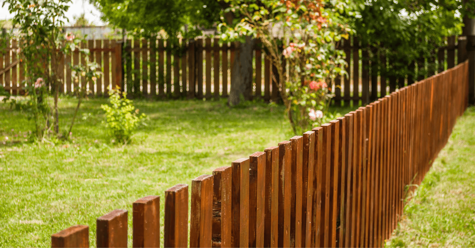 Clôture en bois brun bordant un jardin verdoyant avec pelouse, arbres et fleurs en arrière-plan.