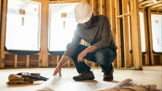 Entrepreneur général portant un casque de sécurité examinant des plans de construction au sol sur un chantier résidentiel.