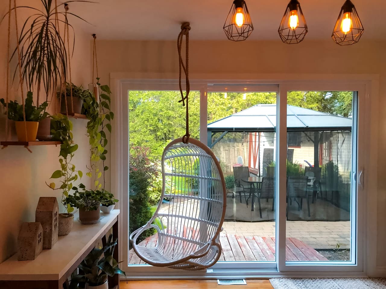Coin détente lumineux avec chaise suspendue, nombreuses plantes d’intérieur, luminaires suspendus et grande porte-patio vitrée donnant sur une terrasse avec gazebo.