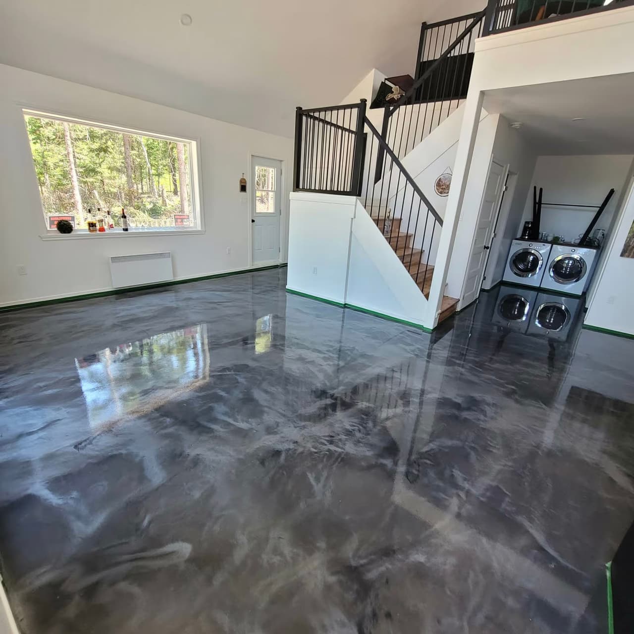 Grande pièce intérieure avec plancher époxy gris lustré, escalier avec rampes noires, mezzanine, fenêtre panoramique et espace buanderie.