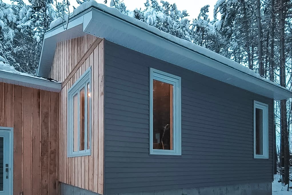 Agrandissement de maison avec façade en bois, fenêtres modernes et fondation en béton, entouré d’une forêt enneigée.