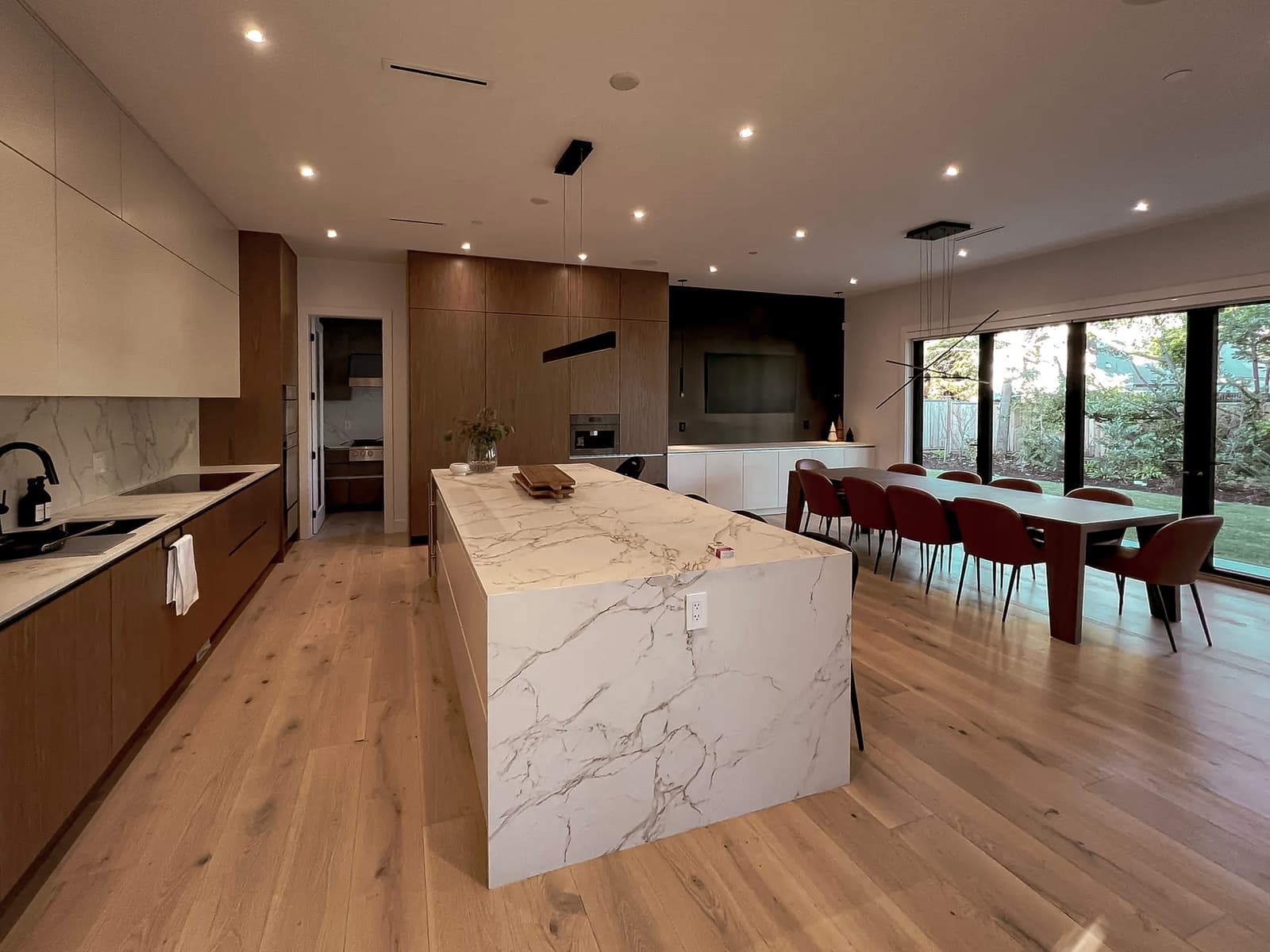 Cuisine moderne ouverte avec îlot en marbre blanc veiné, armoires en bois, sol en bois clair, grande table à manger avec chaises rouges et éclairage suspendu design.