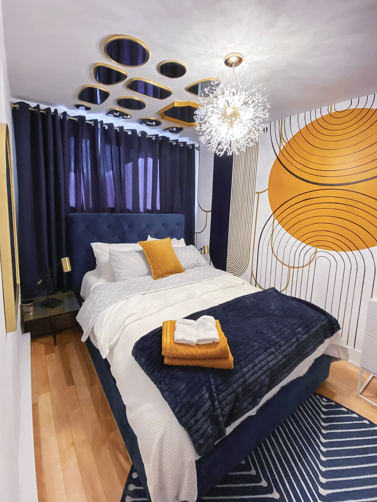 chambre moderne avec lit capitonné bleu, décor mural graphique en jaune et blanc, plafonnier design et rideaux sombres pour une ambiance chaleureuse et élégante.
