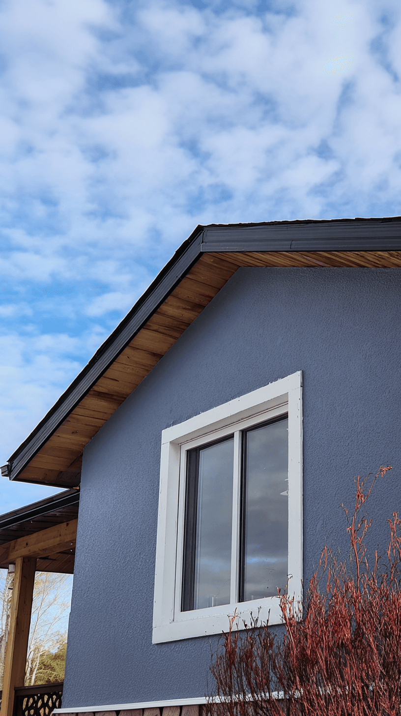 Façade extérieure bleue avec fenêtre blanche, soffites en bois et toiture noire sous un ciel nuageux