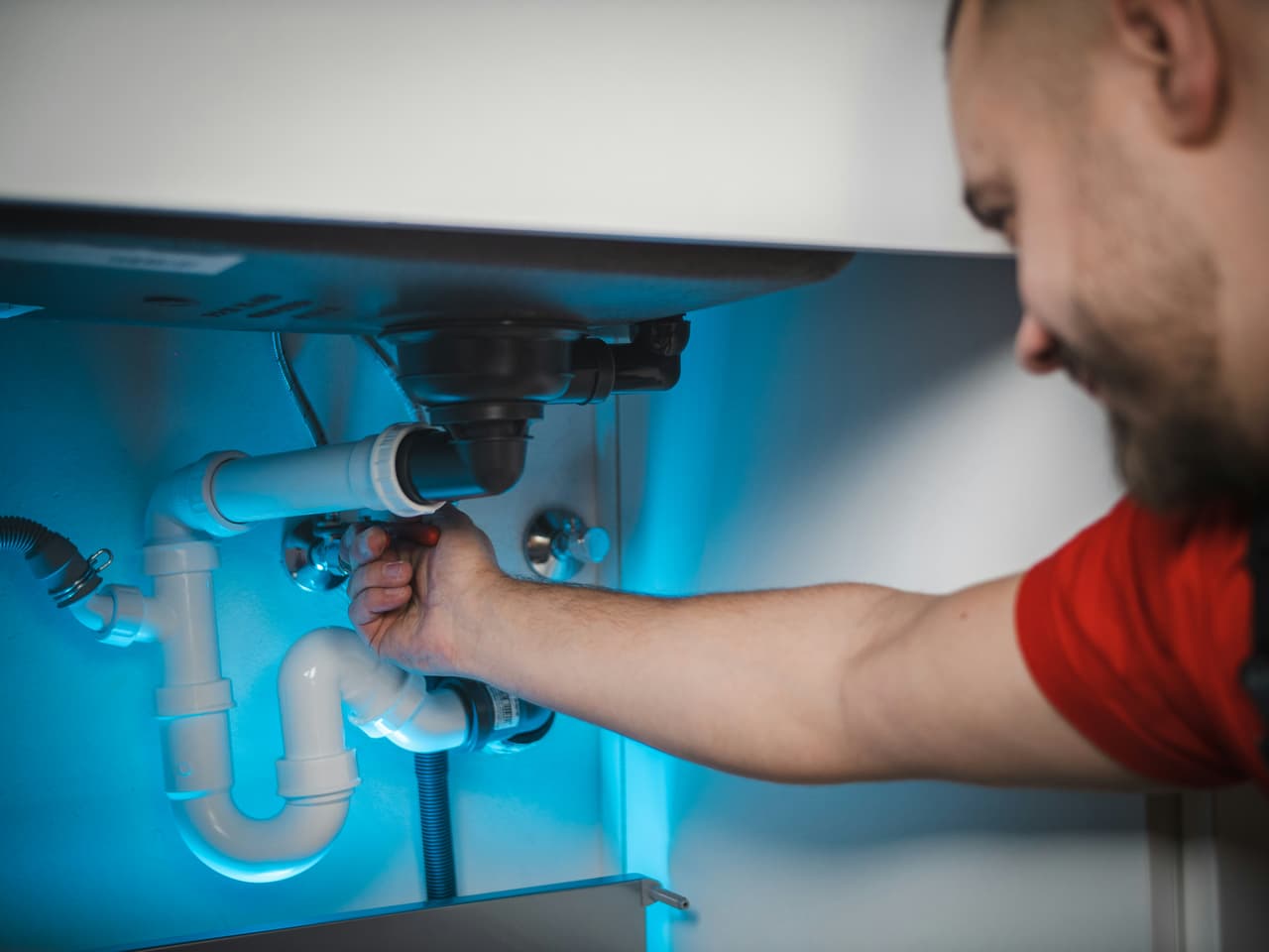 Plombier réparant la tuyauterie sous un évier avec siphon blanc et raccords visibles